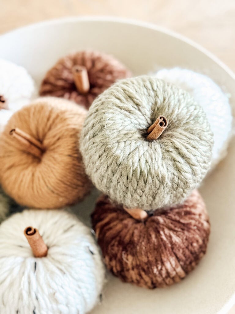 A close-up of decorative pumpkins made from yarn in earthy tones, with cinnamon sticks used as stems, arranged in a white bowl.
