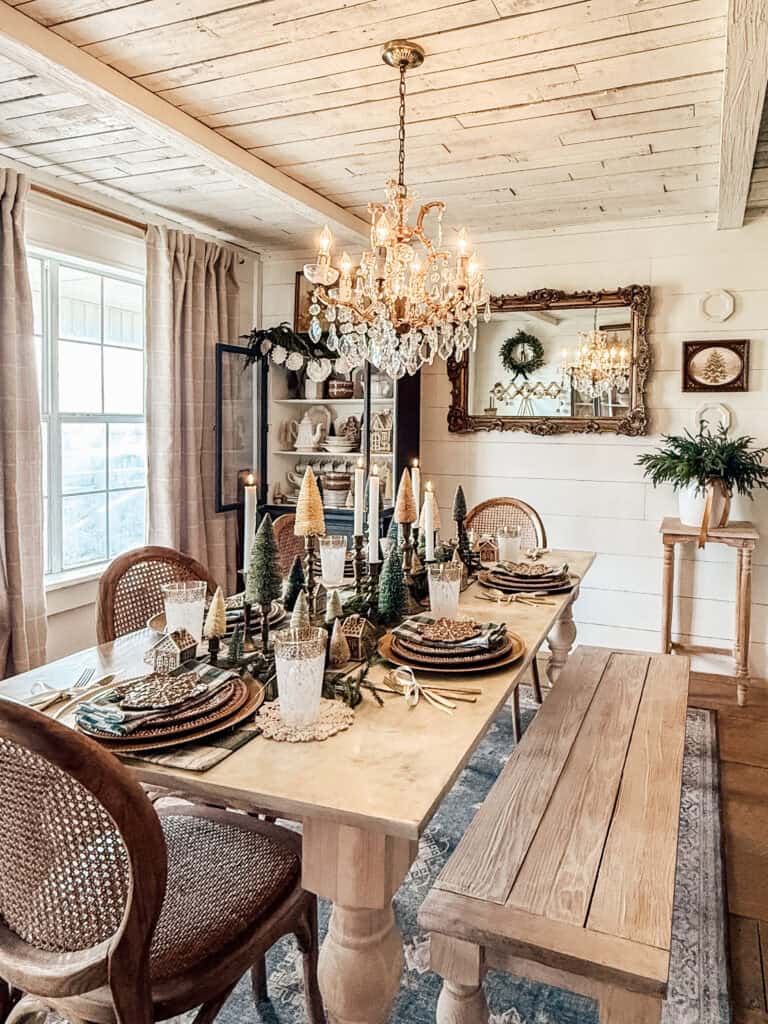 A rustic dining room with a wooden table set for four, decorated with miniature Christmas trees and festive plates. A chandelier hangs above, and a large mirror reflects the cozy, warmly lit space.