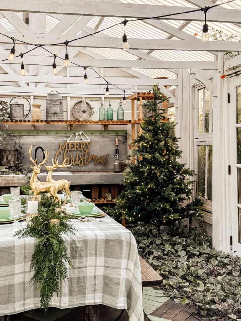 A cozy, rustic room decorated for Christmas with string lights, a Christmas tree with lights, gold reindeer figures on a plaid-covered table, greenery centerpiece, and a Merry Christmas sign on the back wall.