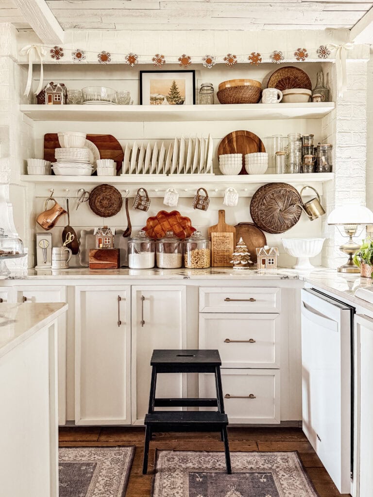 A cozy, rustic kitchen with white cabinets, open shelves displaying dishes, woven baskets, jars, cutting boards, copper utensils, and holiday decor. A small black step stool sits in front of the cabinets.