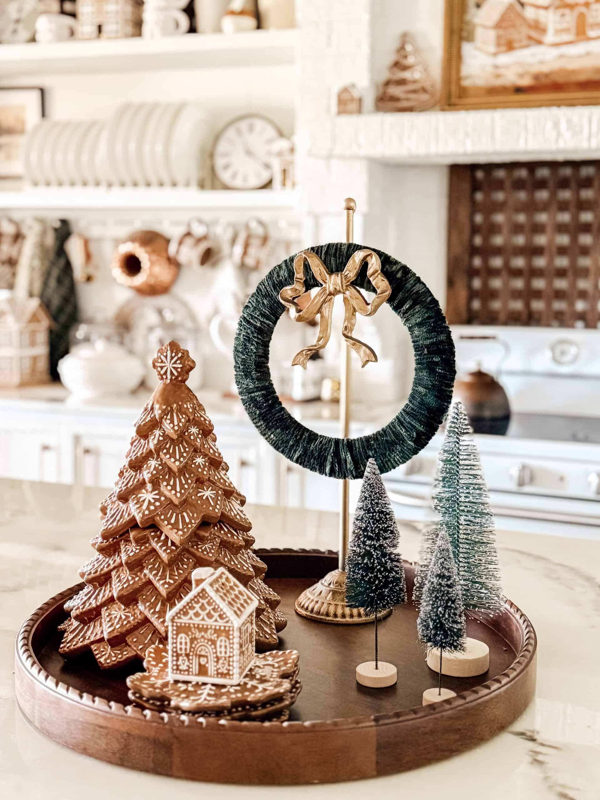A holiday-themed kitchen display features gingerbread trees, a small gingerbread house, mini bottlebrush trees, and a green wreath with a gold bow, all arranged on a round tray atop a marble counter.