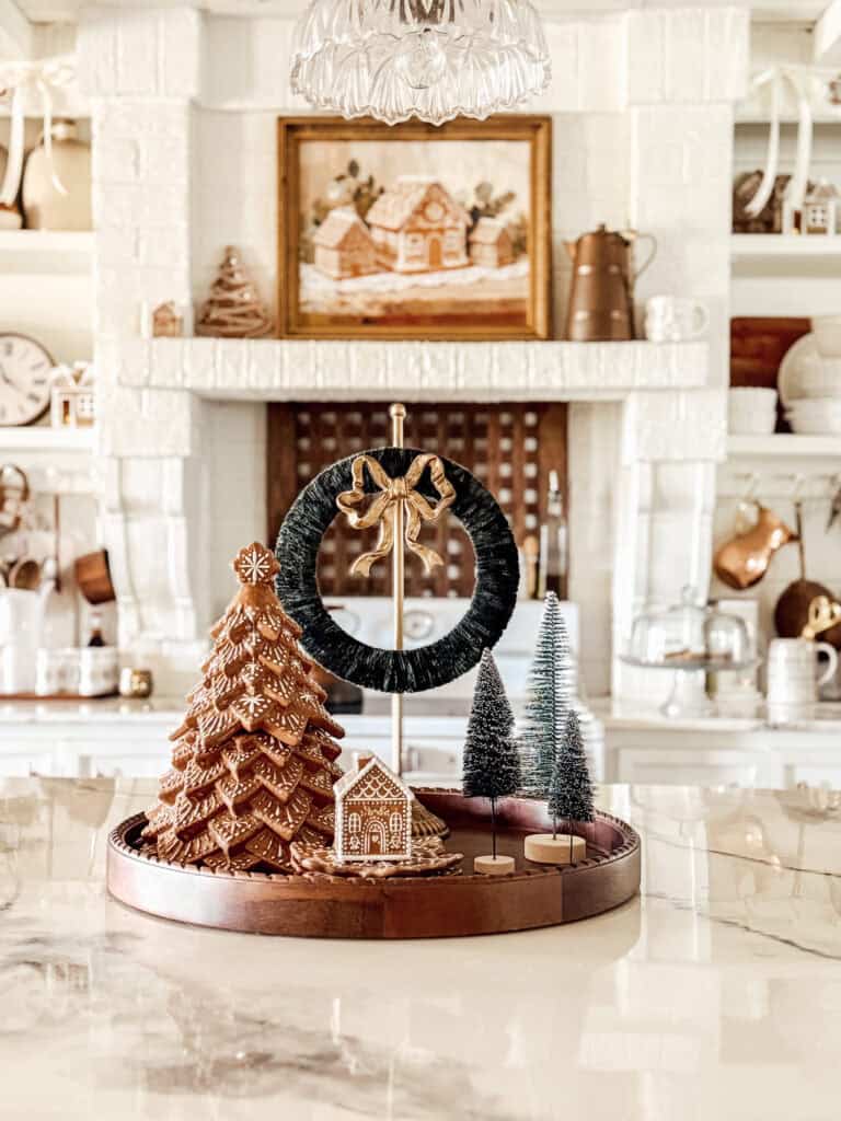 A decorative tray on a marble countertop holds a gingerbread tree, miniature bottle brush trees, a small gingerbread house, and a green wreath. The background features a white fireplace, shelves, and copper kitchen decor.