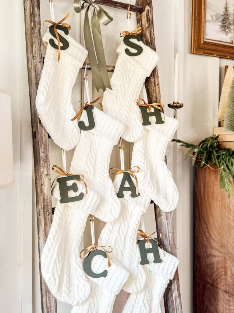Eight white knit Christmas stockings hang on a rustic wooden ladder, each adorned with a large green letter and gold ribbon. The letters spell out SJSHEACH. Greenery and framed art are visible in the background.