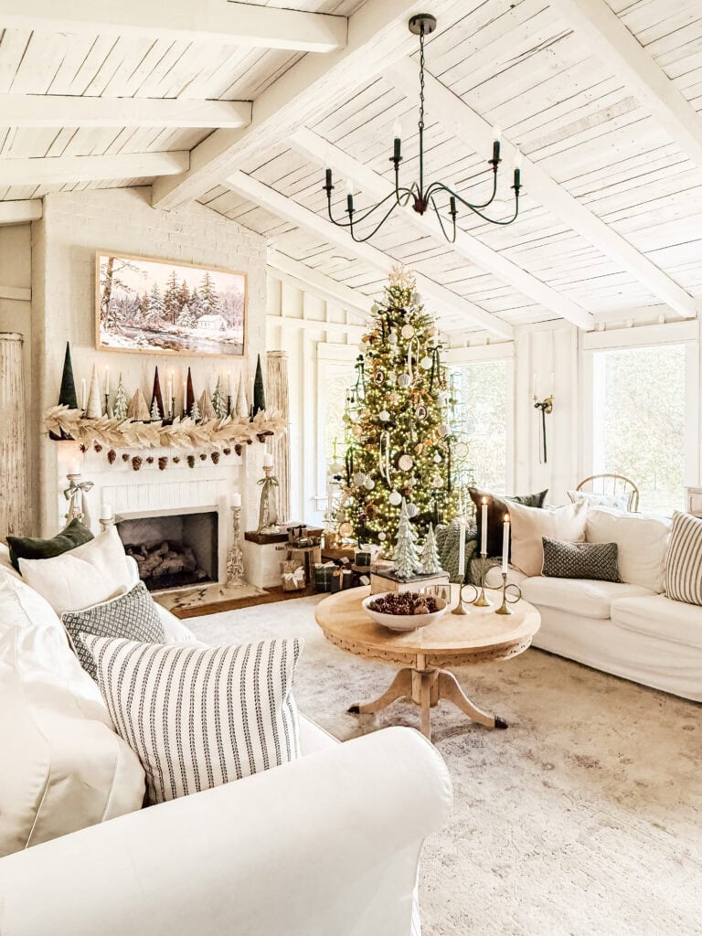 A bright, cozy living room decorated for Christmas with a large tree, white sofas, a wooden coffee table, a garland and mini trees on the mantel, and a chandelier hanging from a white vaulted ceiling.