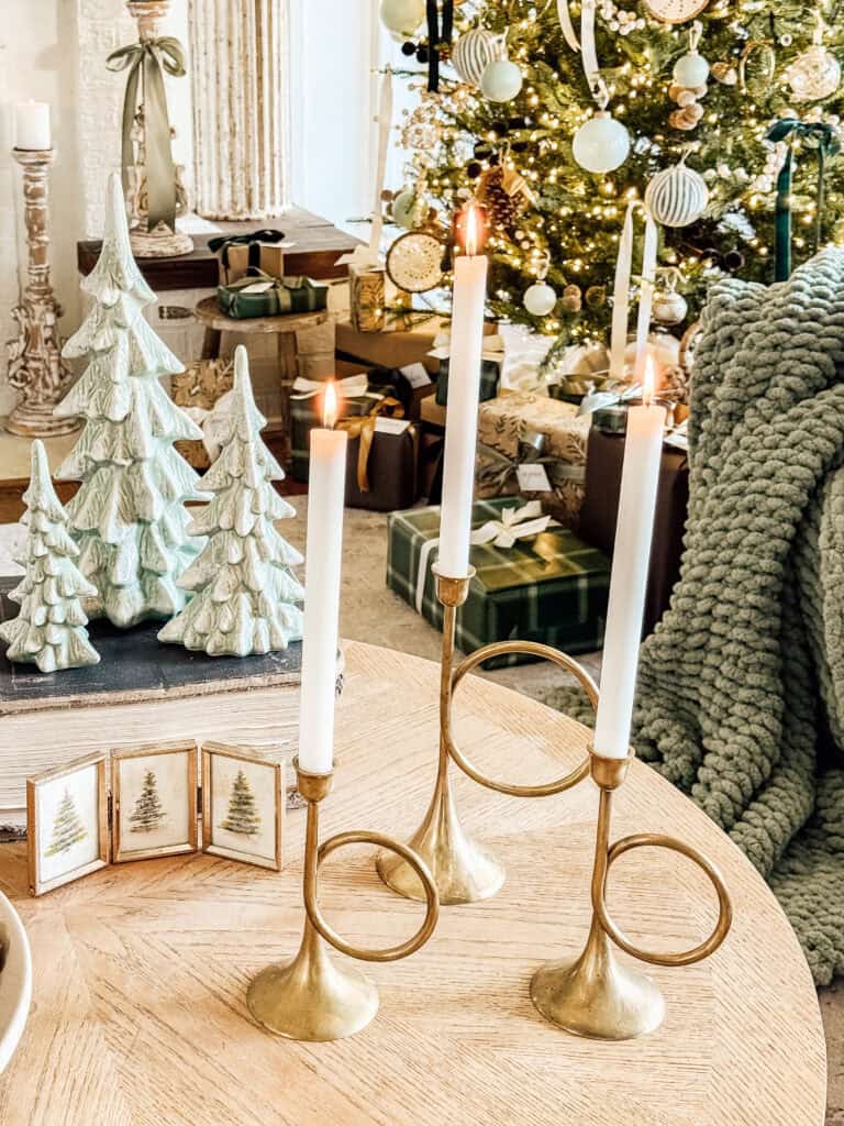 Three lit white taper candles in gold holders sit on a wooden table near ceramic Christmas trees, with a decorated Christmas tree and wrapped presents in the background. Cozy green knit blanket draped on a chair.