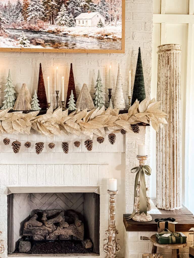 A white brick fireplace mantle decorated with cone-shaped trees, white poinsettias, pinecones, candles, and garland. A snowy landscape painting hangs above, and wrapped gifts sit on a nearby table.