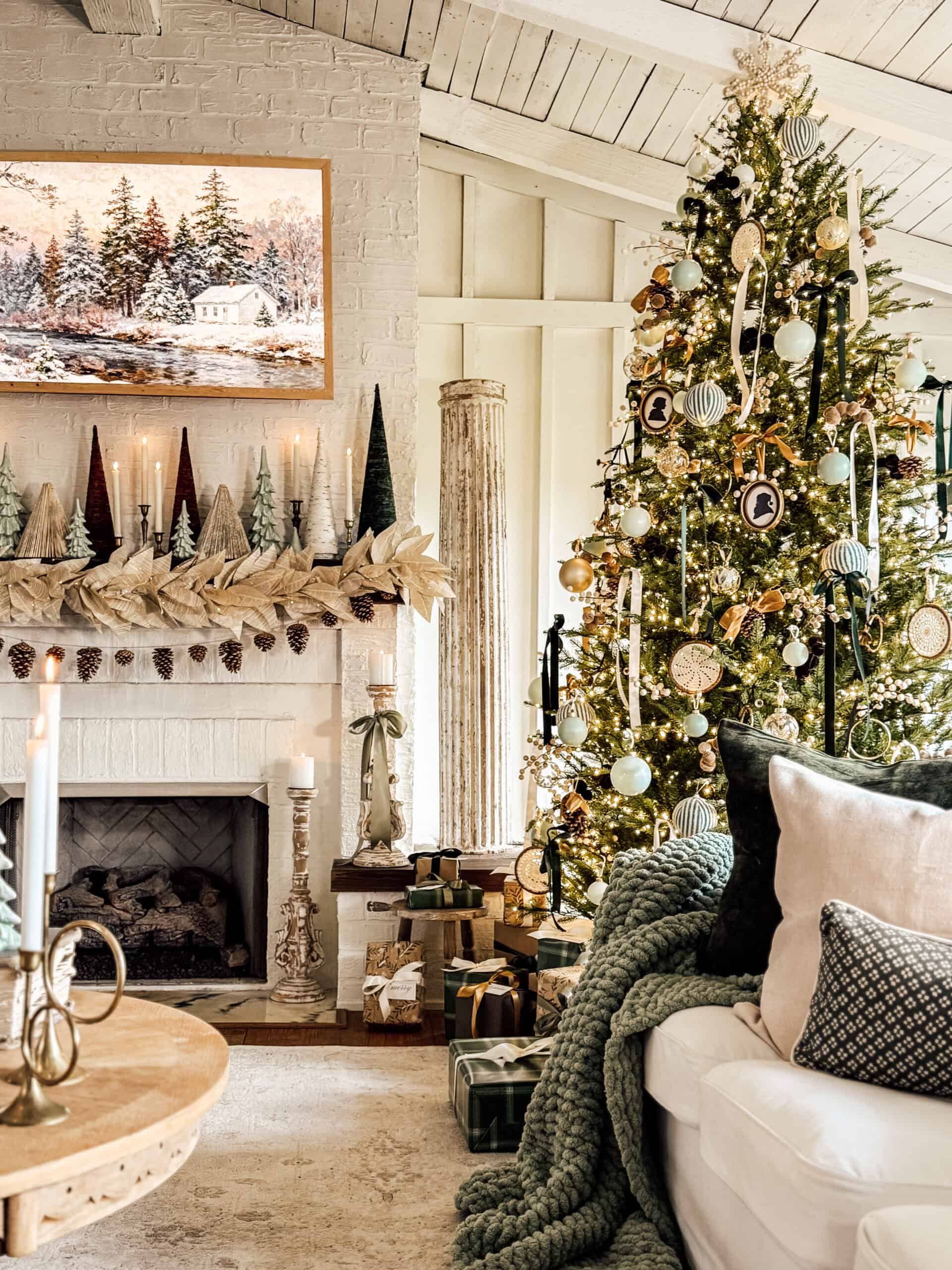 A cozy living room decorated for Christmas with a lit tree, wrapped presents, candles, a garland and tree ornaments on the mantel, and a wintry painting above a white brick fireplace. Plush pillows and blankets are on the sofa.
