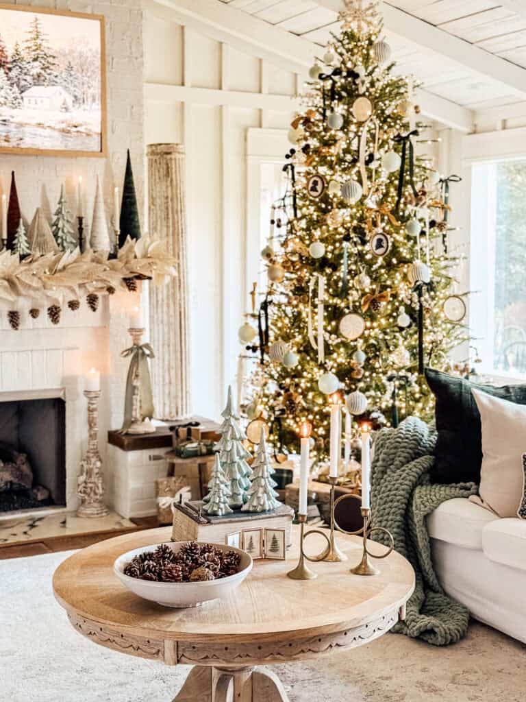 A cozy living room decorated for Christmas with a lit tree adorned with ornaments, a mantel with miniature trees and stockings, a lit fireplace, and a round wooden table holding candles, pine cones, and small decorative trees.