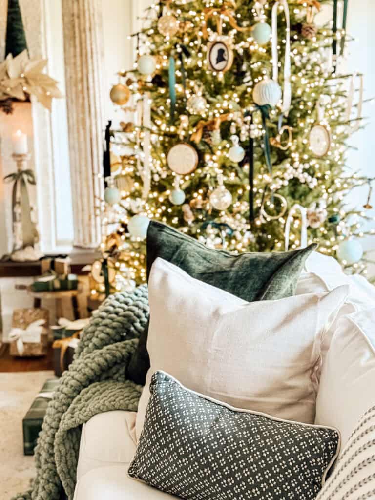 A cozy living room with a white sofa, green and white pillows, a chunky knit blanket, and a decorated Christmas tree with gold, white, and green ornaments, glowing lights, and wrapped presents underneath.
