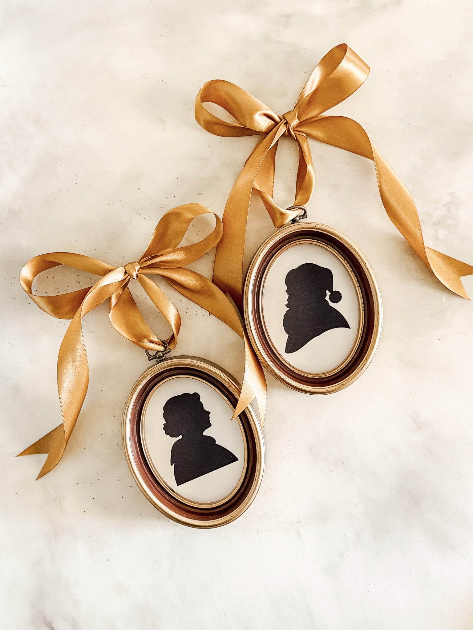Two oval-framed black silhouette portraits hang from gold satin ribbons tied in bows, displayed on a light-colored marble surface—perfect vintage decor inspiration for your Christmas home tour.