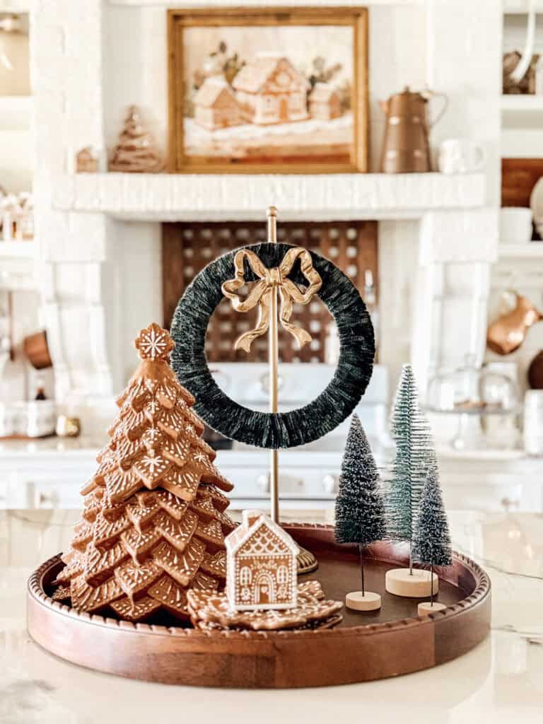 A decorative holiday tray featuring a gingerbread tree, a small gingerbread house, mini bottle brush trees, and a green wreath with a gold bow, set on a kitchen counter with a white brick fireplace in the background.