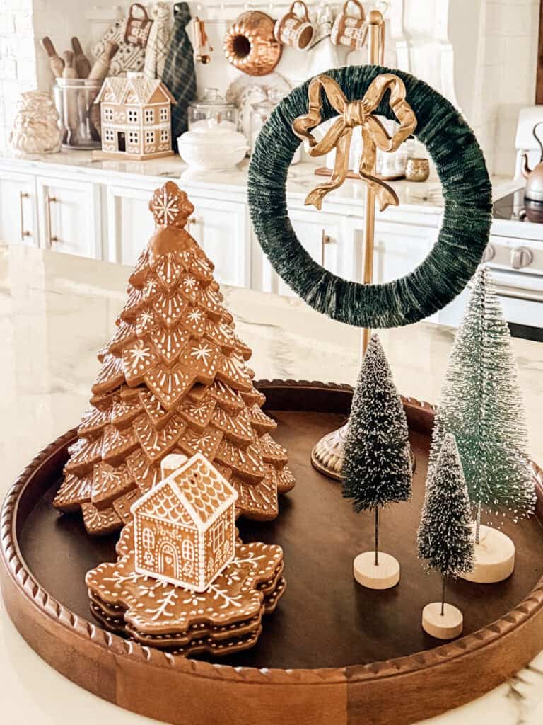 A decorative tray on a kitchen counter features a gingerbread tree, a small gingerbread house, miniature snowy trees, and a green wreath with a gold bow, creating a festive holiday display.