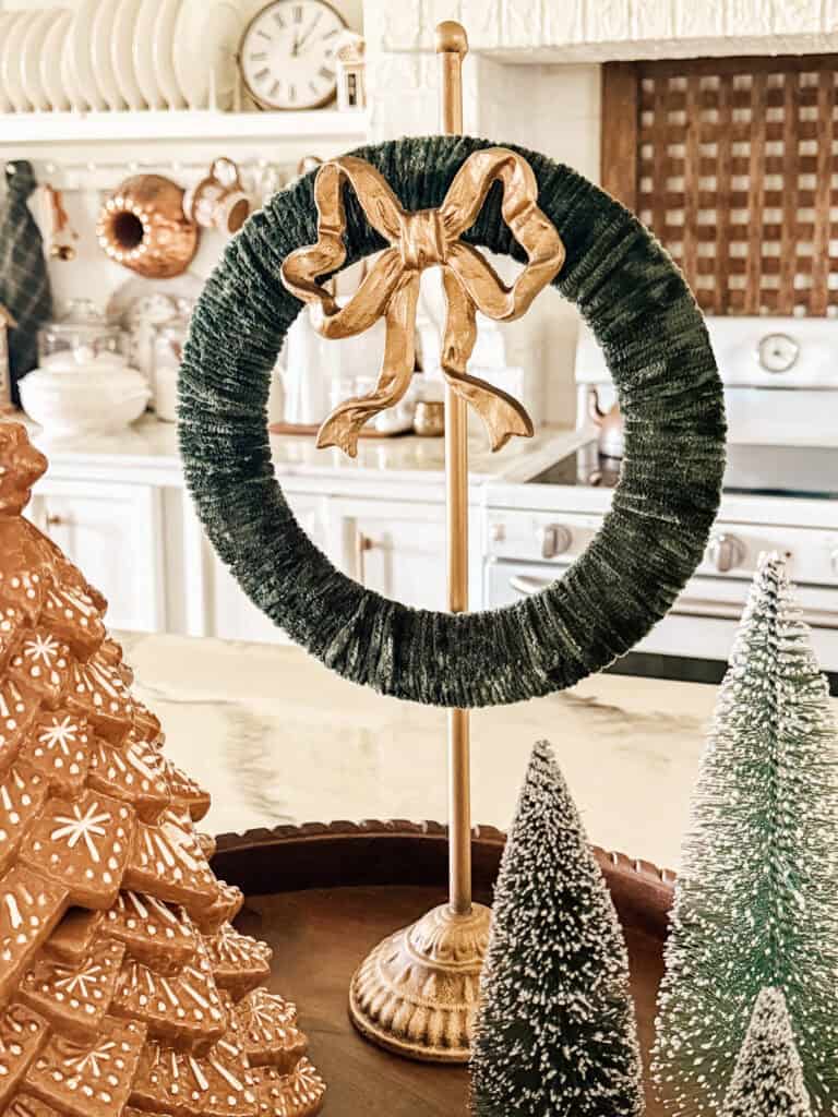 A gold stand holds a green wreath with a gold bow on a kitchen counter, surrounded by a decorative gingerbread-style tree and two bottle brush trees. The kitchen features white cabinetry and copper accents.