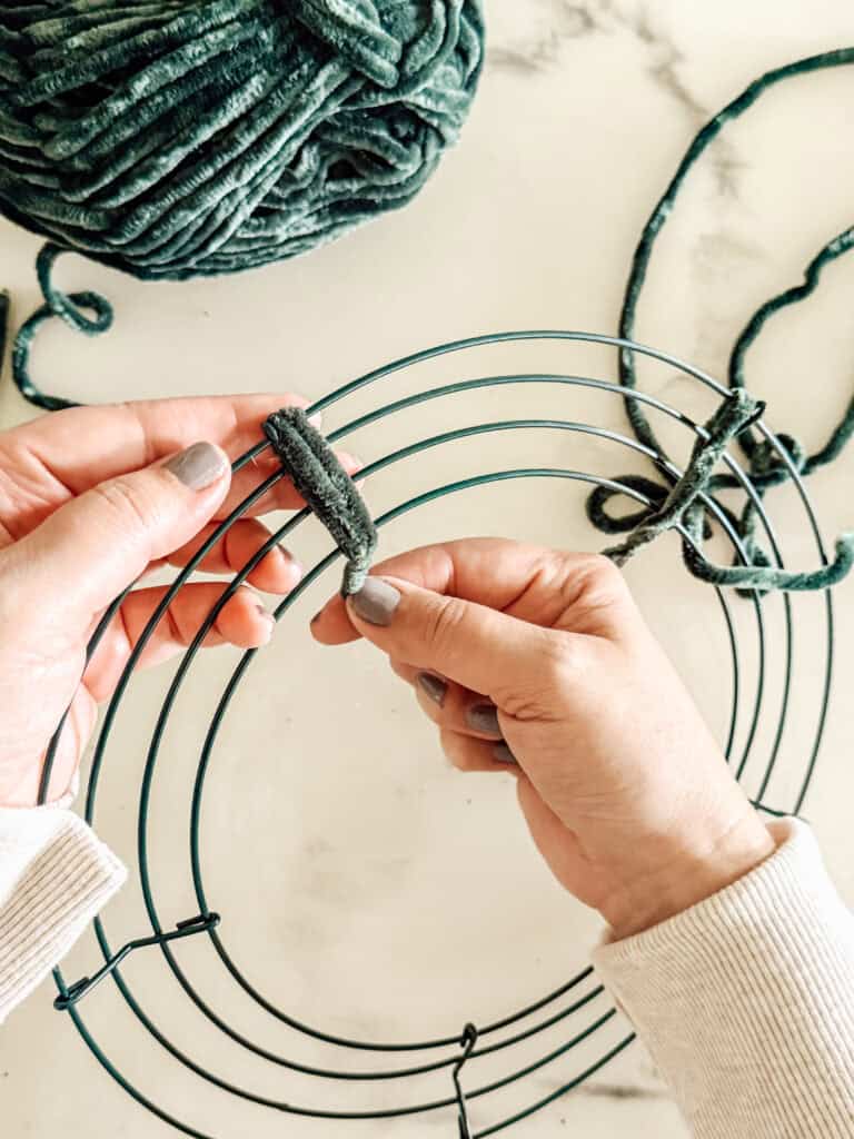 A person’s hands wrap thick green yarn around a metal wire wreath frame on a white surface, with a ball of yarn nearby.