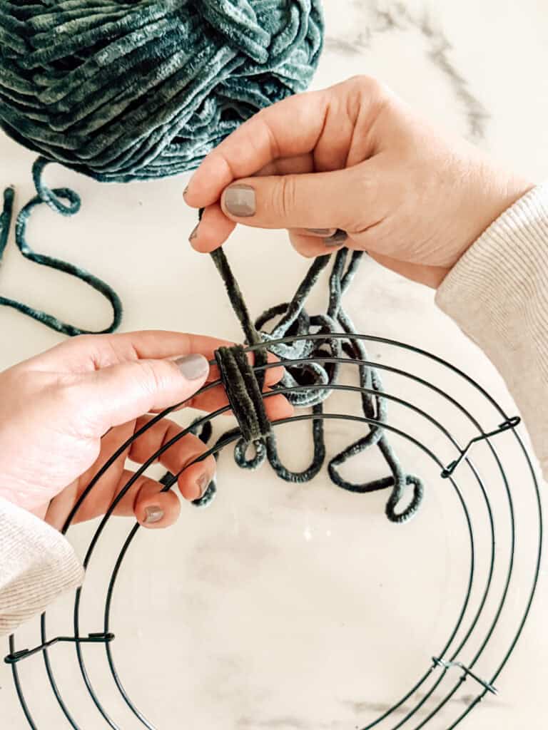 Two hands are tying thick, green yarn onto a round metal wire wreath frame, with a skein of matching yarn nearby on a white surface.
