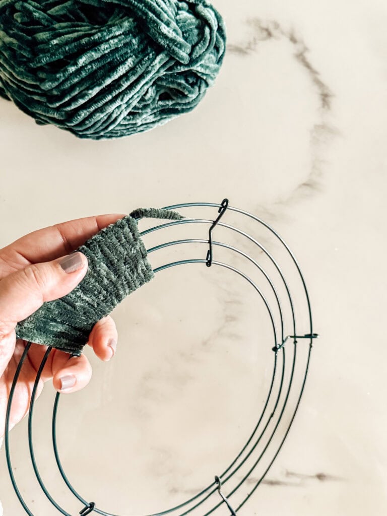 A hand wraps green yarn around a metal wreath frame on a white marble surface, with a ball of matching yarn in the background.