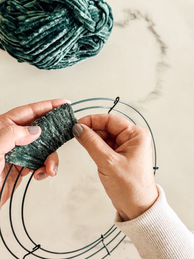 Close-up of hands wrapping thick, dark green yarn around a metal wreath frame on a white surface, with a ball of matching yarn in the background.