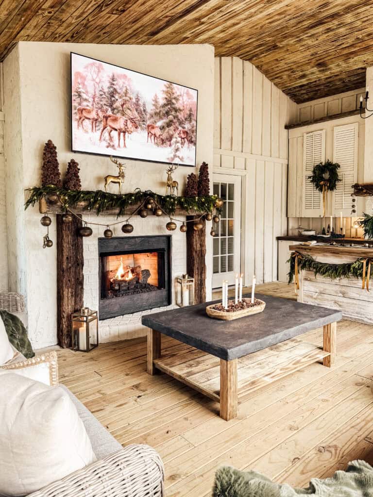 A cozy living room with a rustic wooden ceiling, a lit fireplace decorated with greenery and pinecones, a large TV above showing a winter scene, and a wooden coffee table with candles. Holiday decorations add a festive touch.