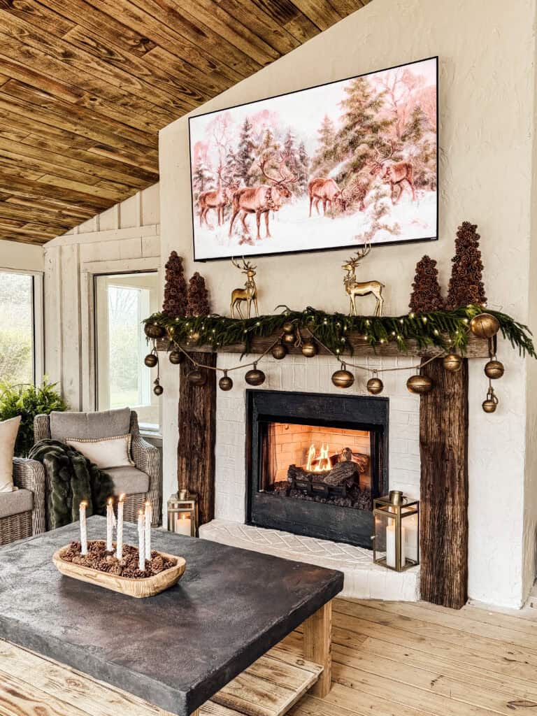 A cozy living room features a lit fireplace with a decorated mantel of pine garlands, bells, and gold reindeer figurines. Above the mantel is a winter woodland painting. A coffee table holds pinecones and candles.