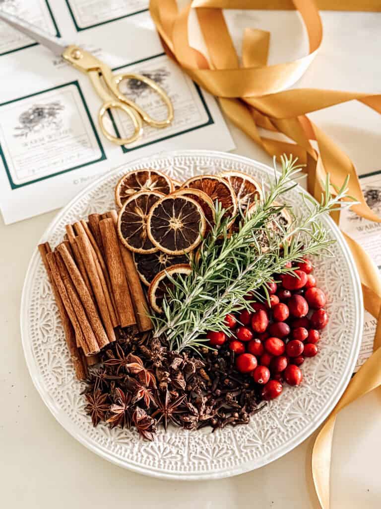 A decorative white plate holds dried orange slices, cinnamon sticks, fresh rosemary, cranberries, star anise, and cloves. Gold scissors, ribbon, and printed labels are nearby on a cream-colored surface.