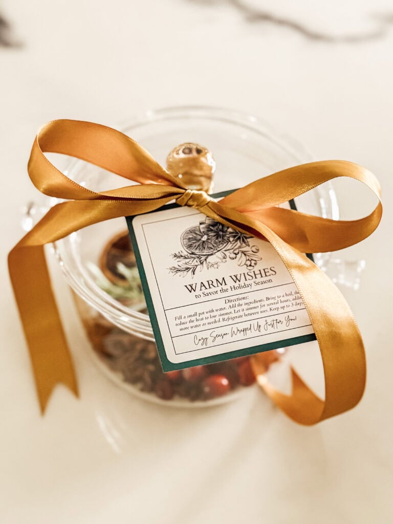 A glass jar filled with treats is topped with a gold ribbon and a decorative tag that reads Warm Wishes and contains a holiday message and instructions; the jar sits on a light surface.
