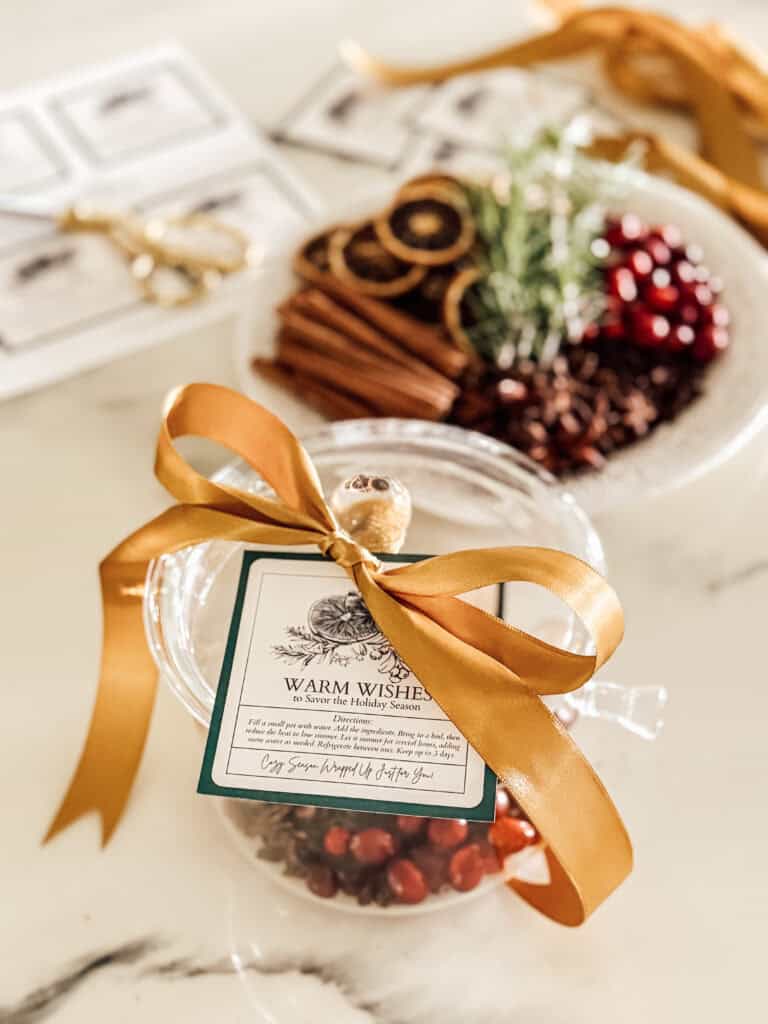 A glass jar filled with cranberries, tied with a gold ribbon and gift tag, sits on a marble counter. In the background, a plate holds dried orange slices, cinnamon sticks, cranberries, and rosemary.