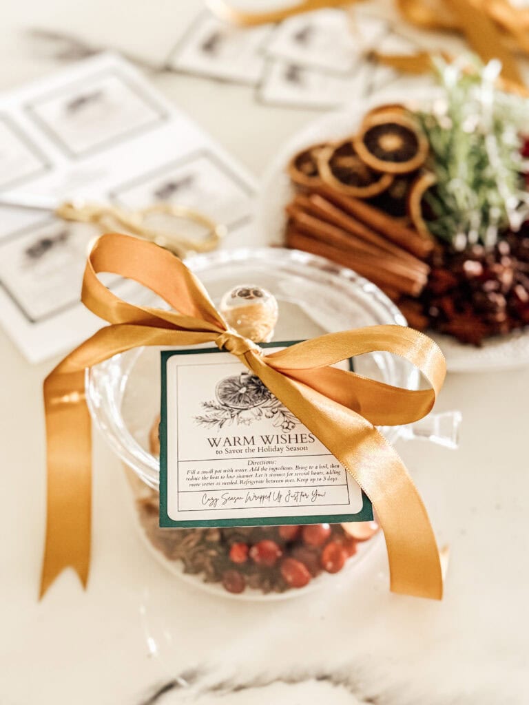 A glass jar filled with cranberries and topped with a gold ribbon and gift tag sits on a table. In the background, there is a plate with dried orange slices, cinnamon sticks, and greenery.