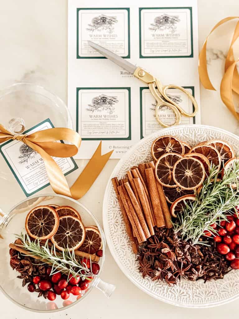 A white plate holds dried orange slices, cinnamon sticks, rosemary, cranberries, and star anise. Nearby are scissors, gold ribbon, glass jars, and printed Warm Wishes gift tags on a marble surface.