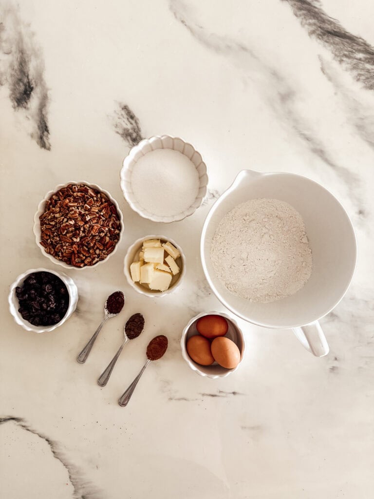Overhead view of baking ingredients on a marble surface: a bowl of flour, a bowl of sugar, cubed butter, eggs, chopped pecans, dried fruit, and three teaspoons with spices.