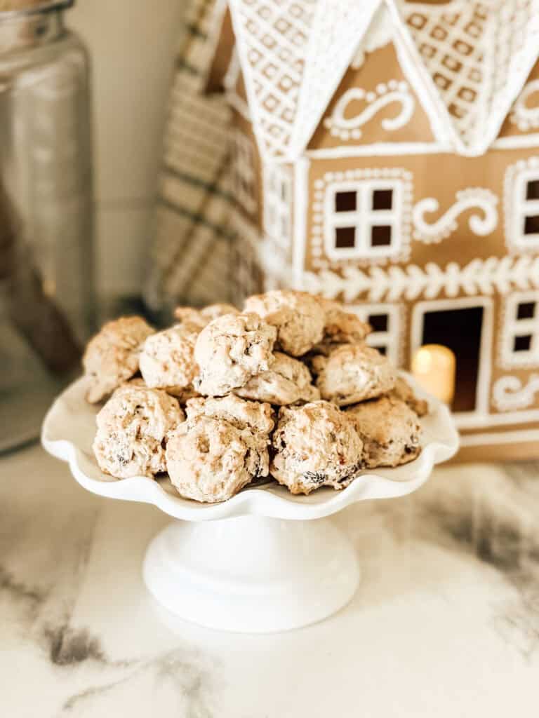 A white pedestal plate holds a pile of cookies in front of a decorative gingerbread house with white icing details on a marble countertop.