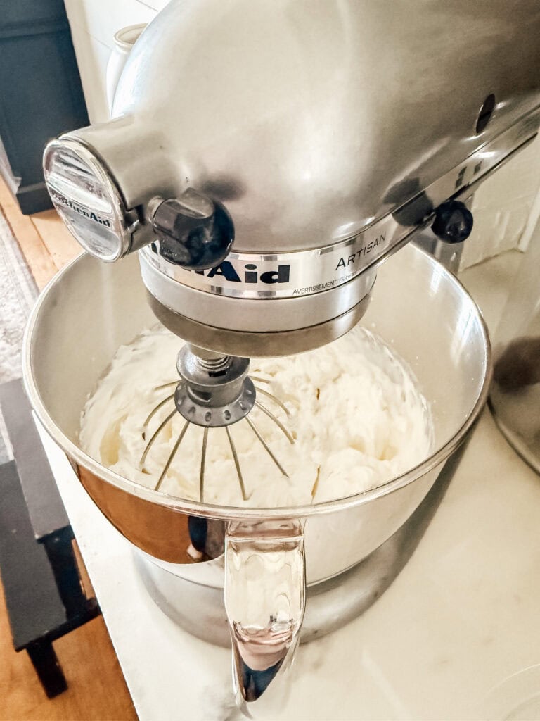 A silver KitchenAid stand mixer with a whisk attachment is whipping a creamy mixture in a metal bowl on a light-colored countertop.