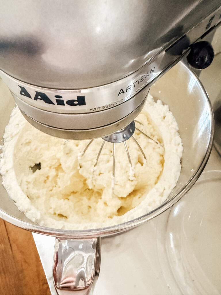 A stand mixer with a wire whisk attachment is mixing a creamy white batter in a stainless steel bowl on a kitchen counter.