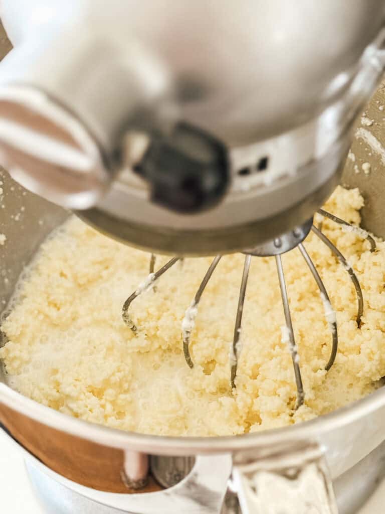 A close-up of a stand mixer with a whisk attachment blending a pale, crumbly mixture in a metal bowl. The mixture appears to be in the early stages of baking preparation.