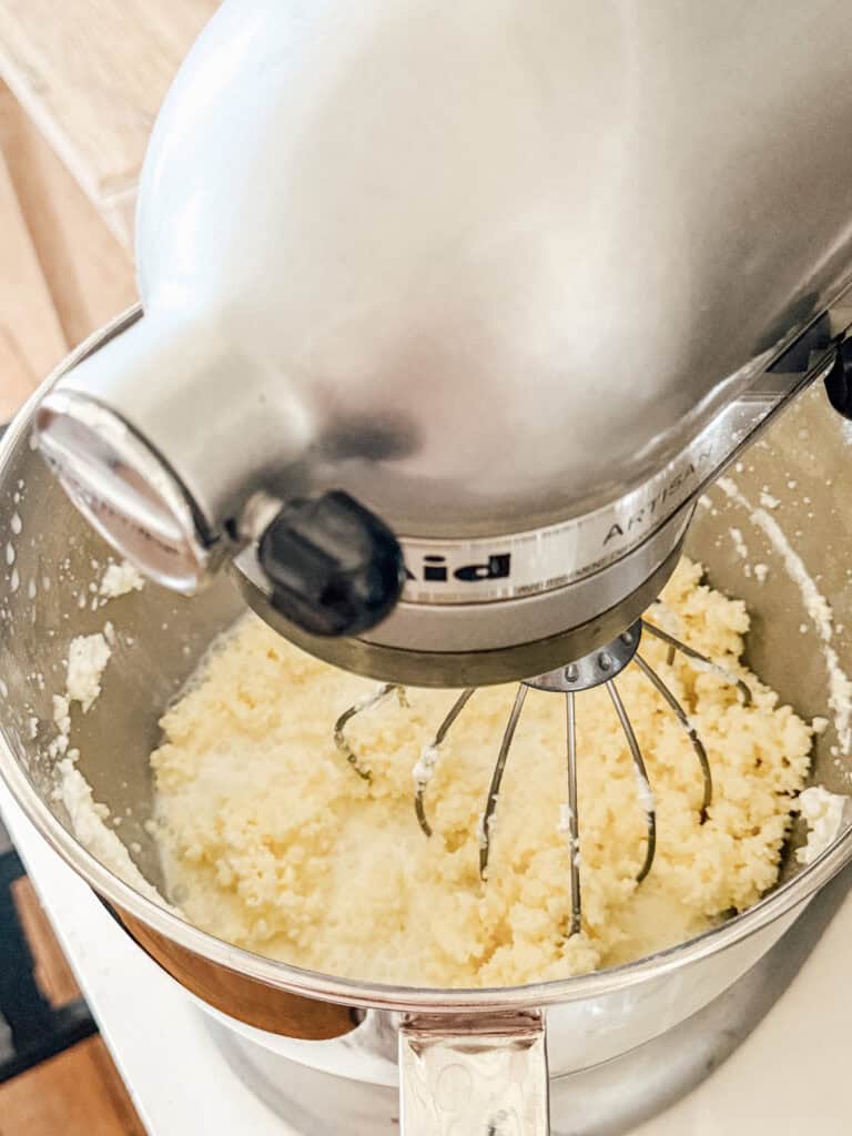 A stand mixer with a wire whisk attachment is whipping a yellow, crumbly mixture in a metal bowl, likely in the process of making butter or creaming ingredients.