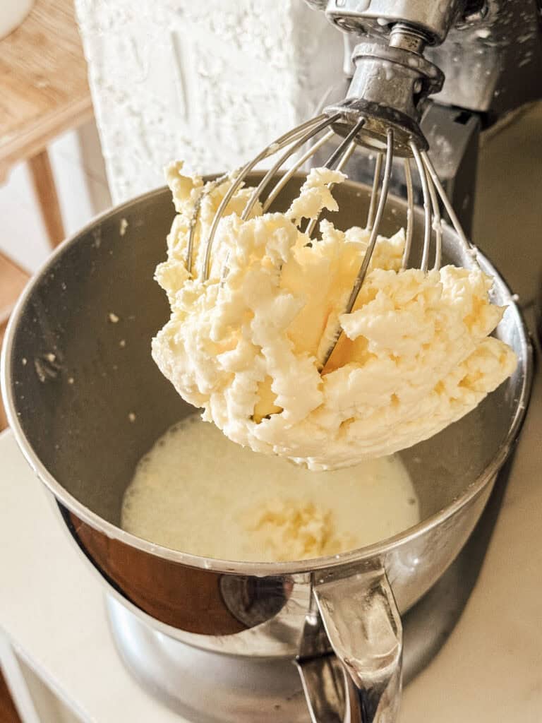 A stand mixer with a whisk attachment holds freshly made butter, while liquid buttermilk remains in the mixing bowl below. The mixer and bowl are situated on a kitchen counter.