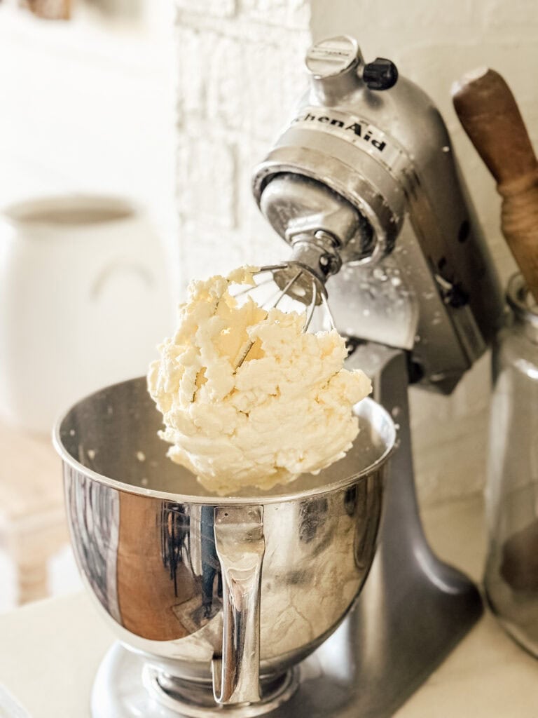 A stand mixer with a whisk attachment holds a large amount of creamy, whipped mixture. The metal mixing bowl is partially filled, and kitchen items are visible in the background.