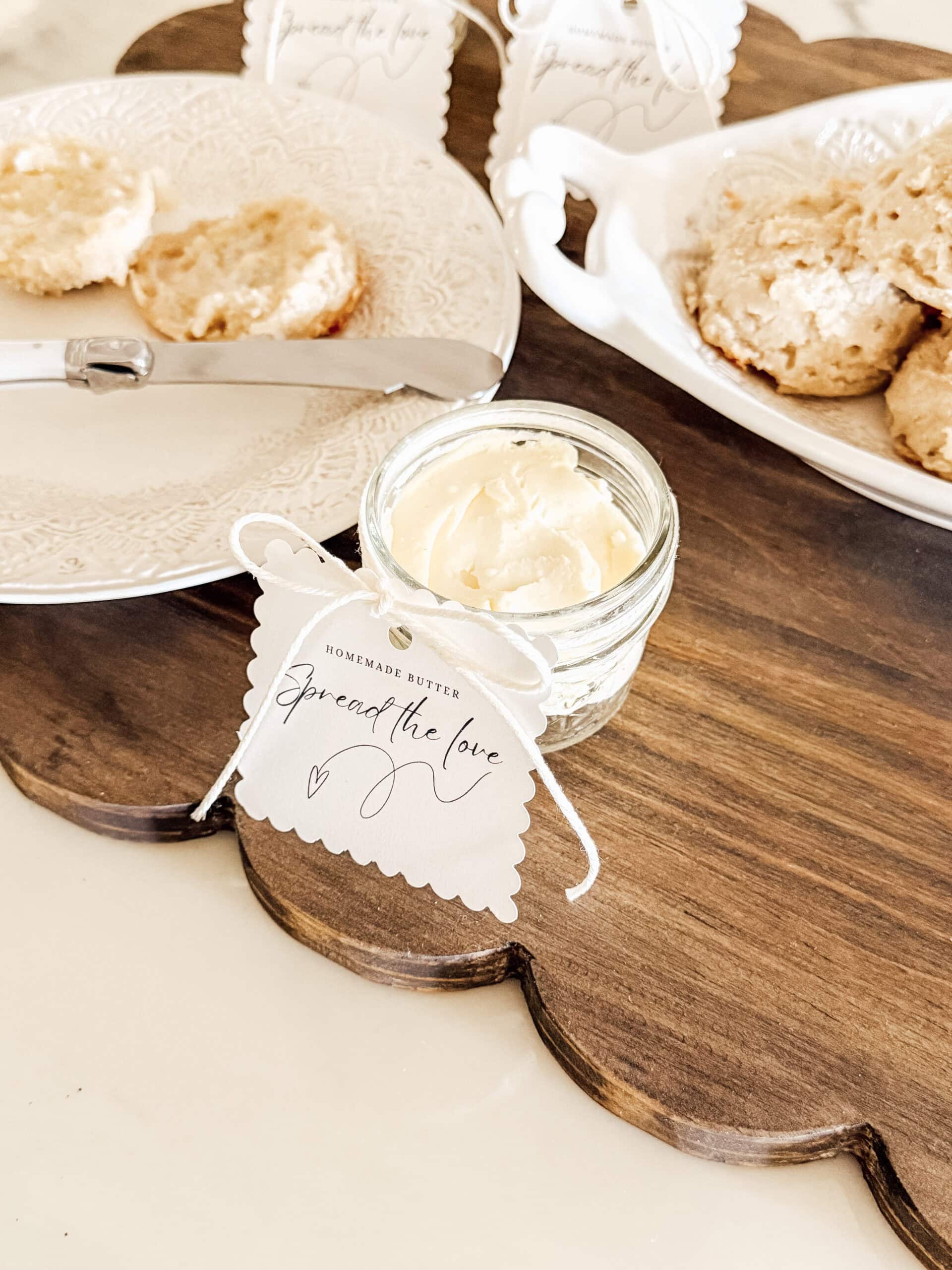 A small jar of homemade butter with a tag that reads Spread the love sits on a wooden board next to a plate of biscuits and a butter knife.