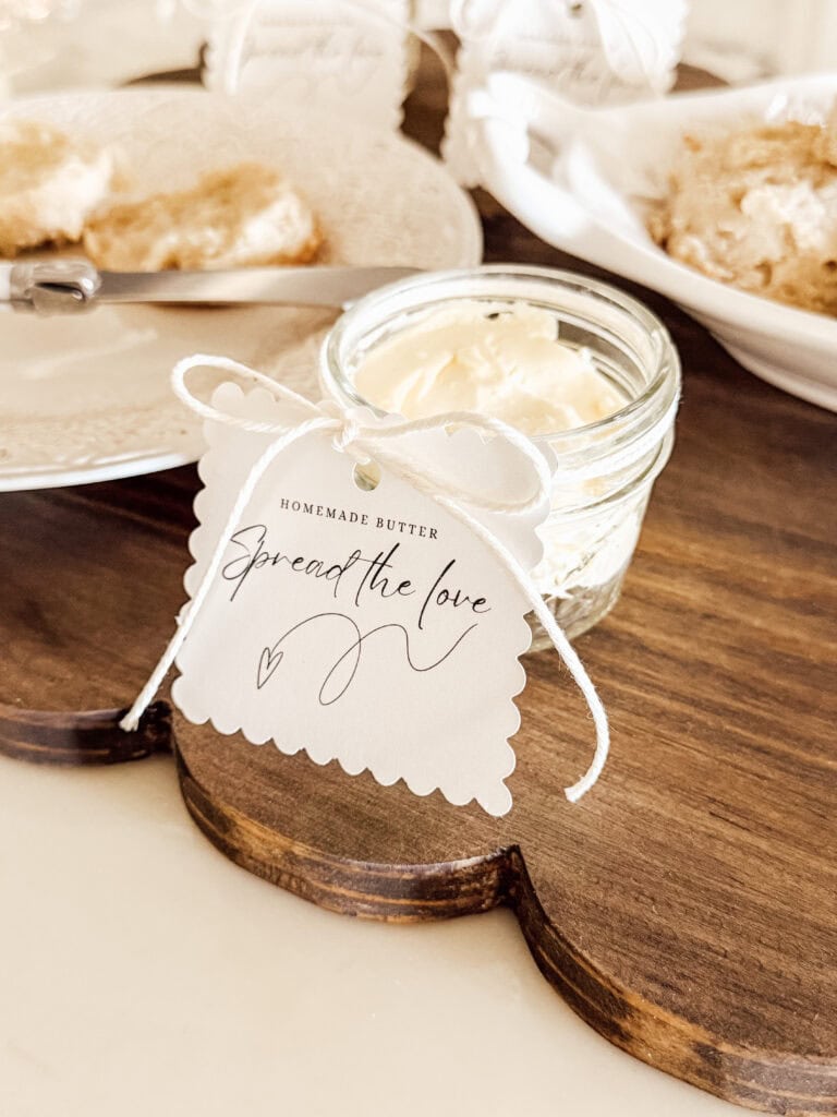 A small jar of homemade butter with a decorative tag reading Spread the Love tied to it, placed on a wooden board next to plates with scones or biscuits.