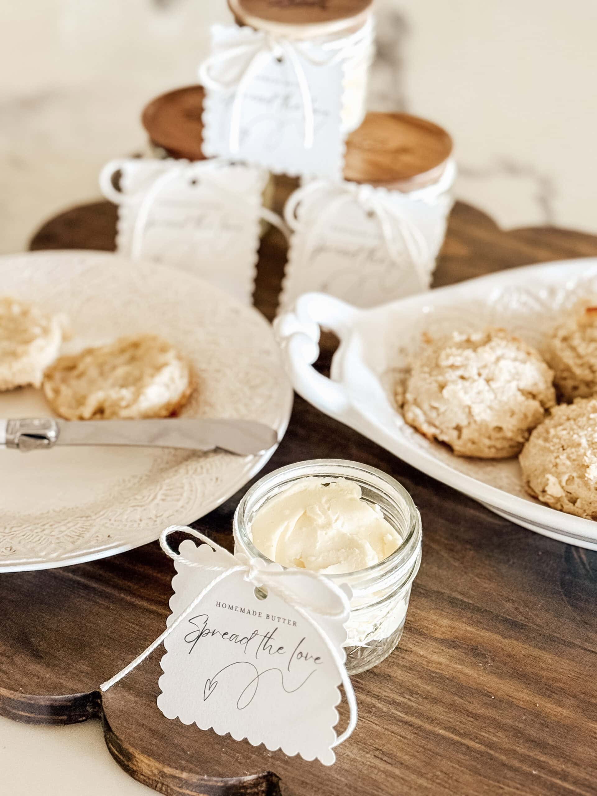 A small jar of homemade butter with a tag reading Spread the love sits on a wooden tray next to biscuits, a knife, and more jars with similar tags and wooden lids in the background.