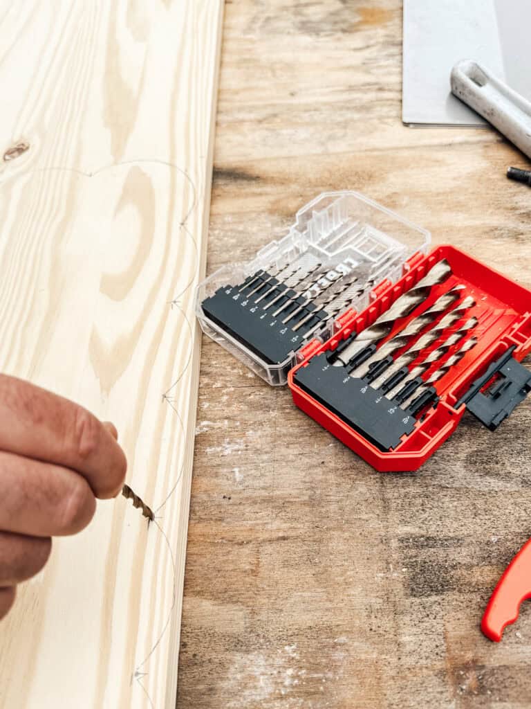 A person holds a small drill bit near a wooden plank with pencil markings. An open red and black drill bit set lies on a workbench beside the wood.