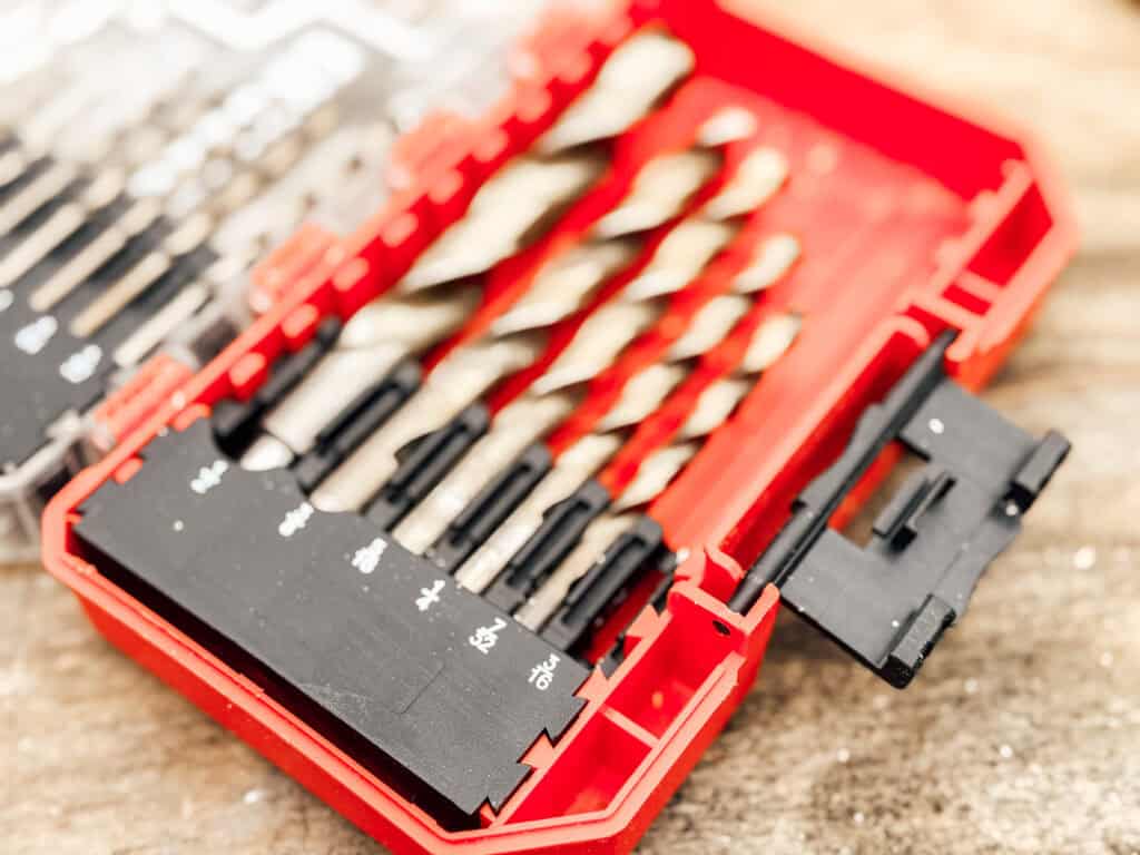 A close-up of a red plastic case containing various drill bits, neatly arranged in slots, with size markings visible on the black interior panel. The case is open and resting on a wooden surface.