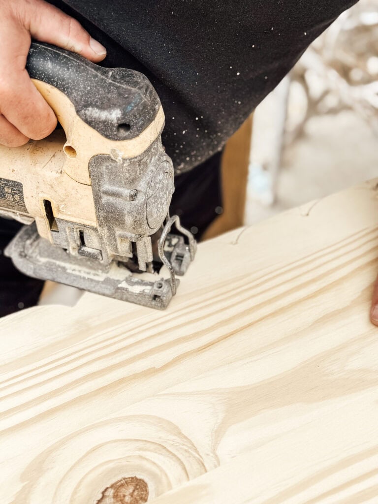 A person uses a handheld jigsaw to cut through a piece of light-colored wood, guiding the saw along a curved line. The person’s hand and part of the tool are visible.