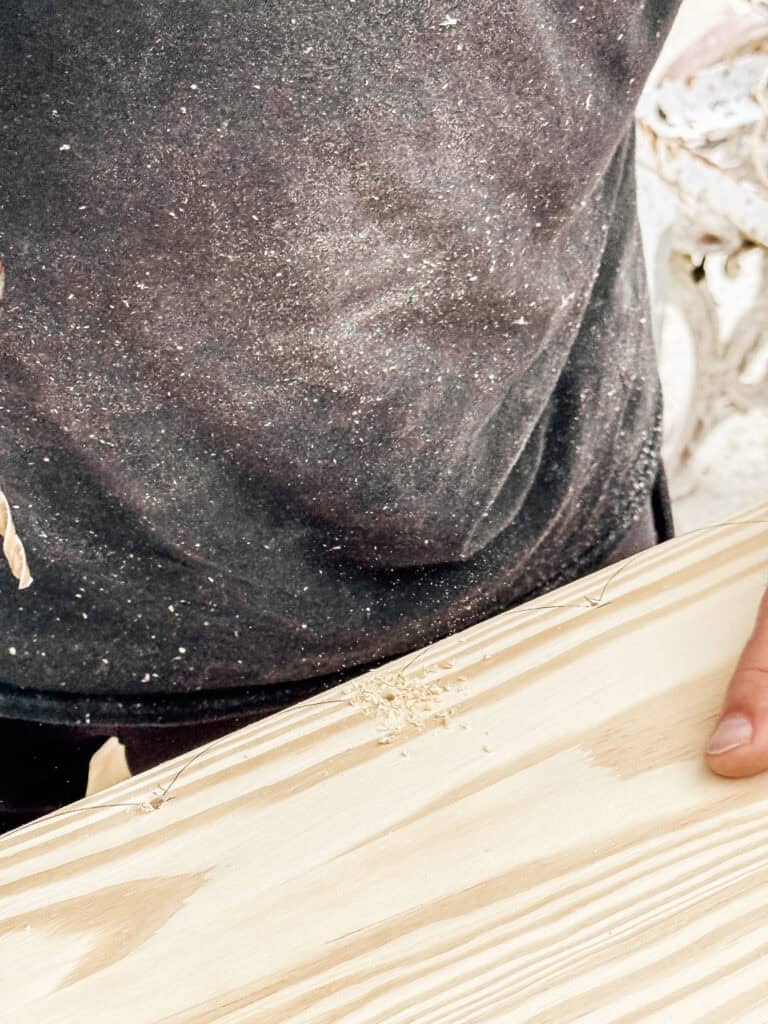A person wearing a dark, sawdust-covered shirt holds a light-colored wooden plank with pencil markings and wood shavings on it.