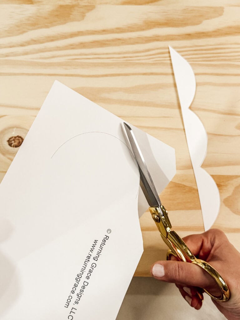 A hand uses gold scissors to cut a curved shape out of white paper on a light wood surface. Another scalloped-cut piece of paper lies nearby.