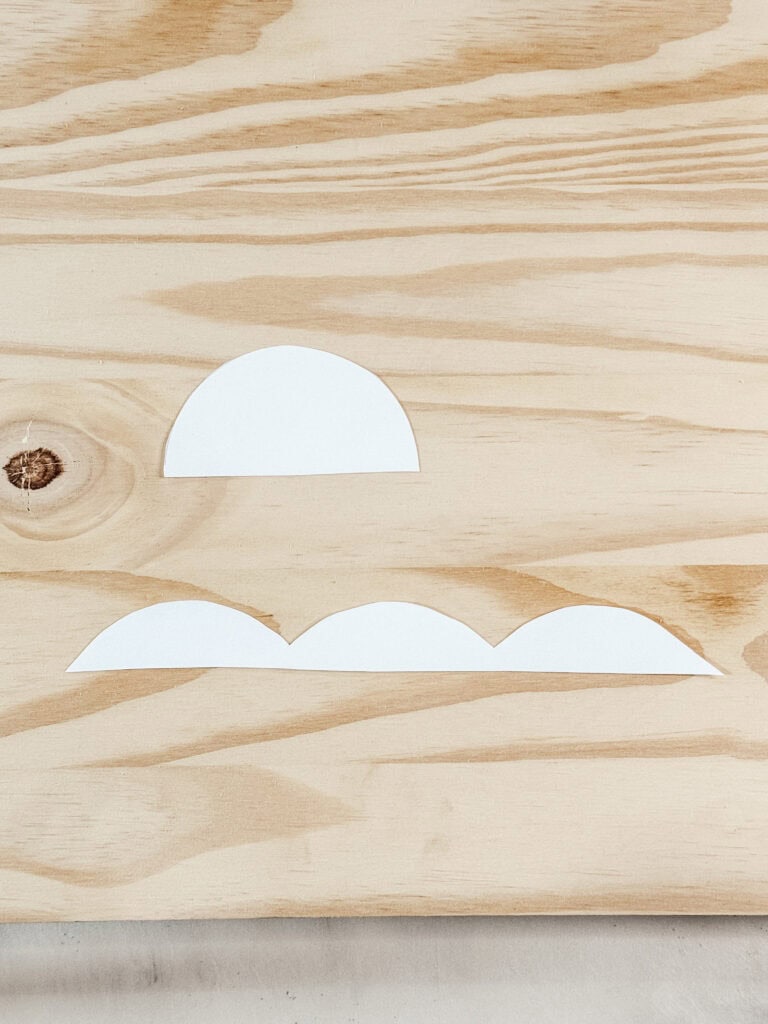 A wooden surface with a white paper semicircle above three white paper scallop shapes arranged in a row. The wood has visible natural grain patterns.