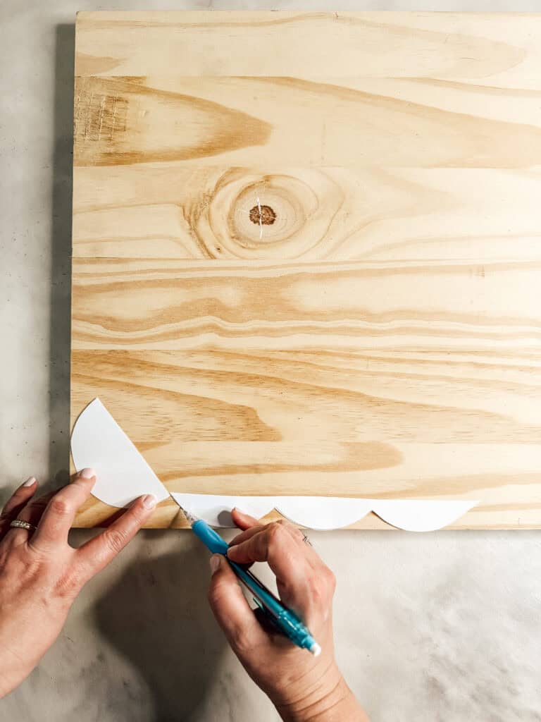 A person traces a scalloped pattern onto a wooden board using a piece of white paper and a blue pencil.