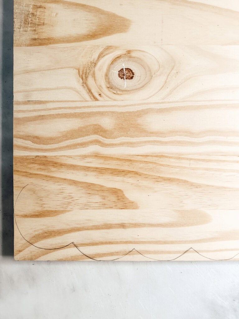 A piece of light-colored wood with visible grain patterns and a knot, resting on a white surface. Pencil-drawn scallop shapes are sketched along the bottom edge of the wood.