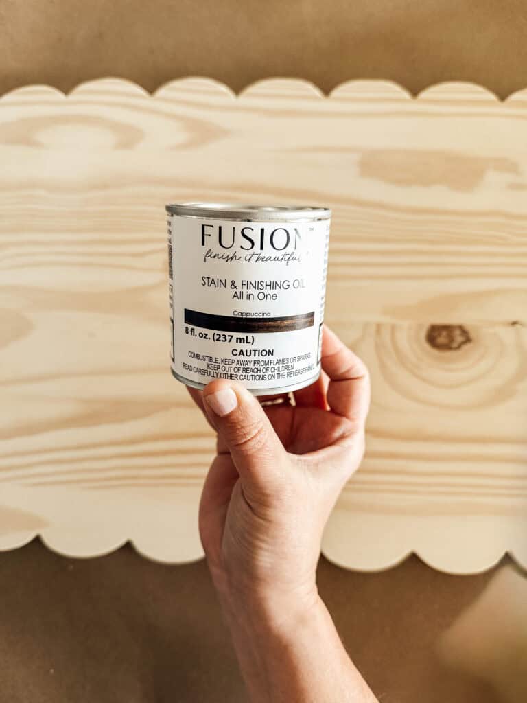 A hand holds a can of Fusion brand stain and finishing oil in front of a light-colored wooden board with a scalloped edge.