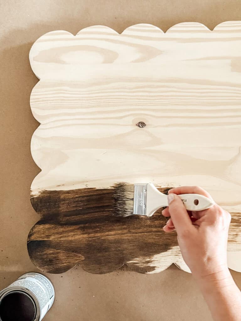 A hand holds a paintbrush, applying dark wood stain to the bottom edge of a scalloped, light wood board. An open can of stain is visible in the lower left corner.