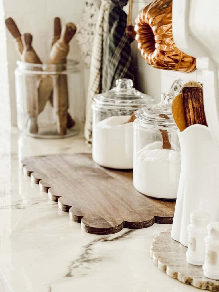A kitchen counter with glass jars of flour and sugar, wooden utensils in a glass jar, rolling pins, a wooden cutting board, and white kitchen tools in a holder, all arranged on a marble surface.